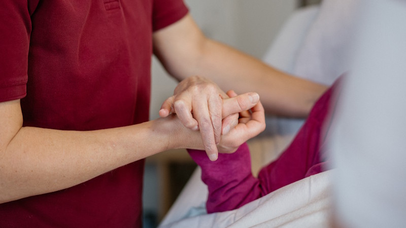 Pflegekraft unterstützt eine pflegebedürftige Person im Bett, hält ihre Hand und begleitet sie einfühlsam im Rahmen der Grundpflege.